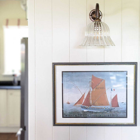 Bodium Small Ribbed glass shade wall light with antique brass finish above framed sailboat artwork in a modern interior.