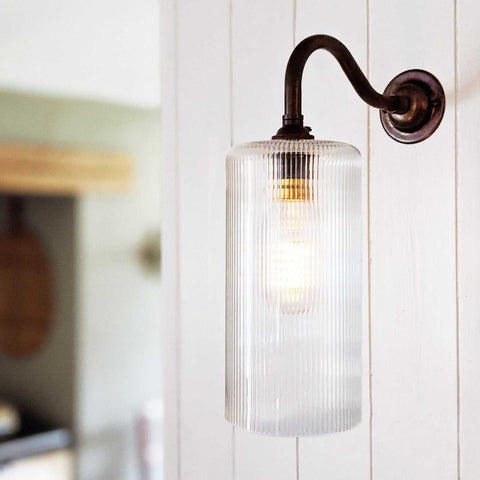 Brook small ribbed glass shade wall light with bronze curved arm mounted on white wooden wall, softly diffusing warm light