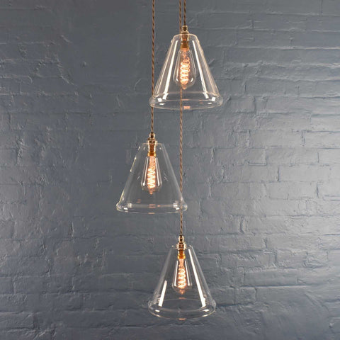 Rye medium clear glass shade cluster pendant light with three hand-blown conical glass shades and adjustable cords against a textured gray brick wall background.