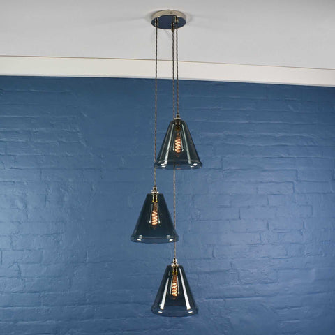Rye medium smoked glass shade cluster pendant light with three conical smoked glass shades and brushed metal fittings hanging against a blue brick wall